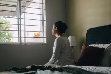 Woman looking out a window while sitting on her bed